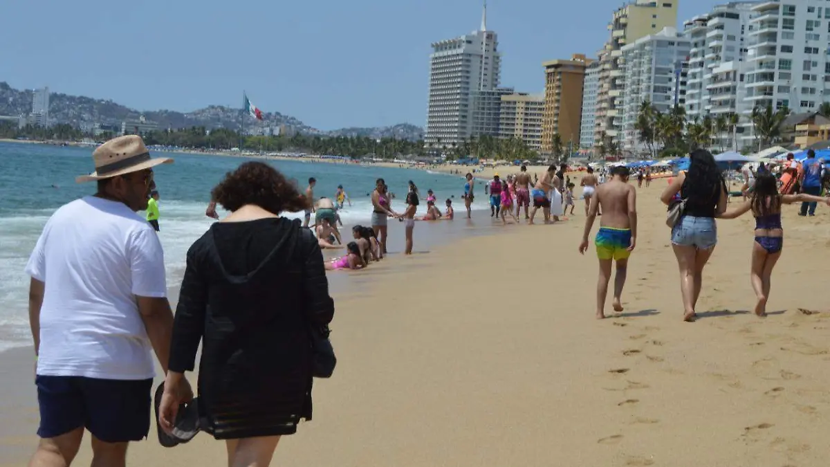 Una pareja en primer plano camina sobre las playas de la zona hotelera de Acapulco entre más paseantes